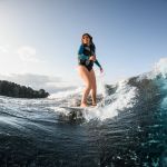 Handsome young woman in swimming vest wakesurfing down the river waves. Beautiful blue sky on the background